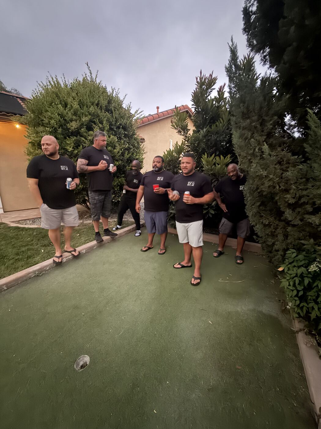Bocce ball in the backyard at dusk — six brothers in SFD tees, drinks in hand