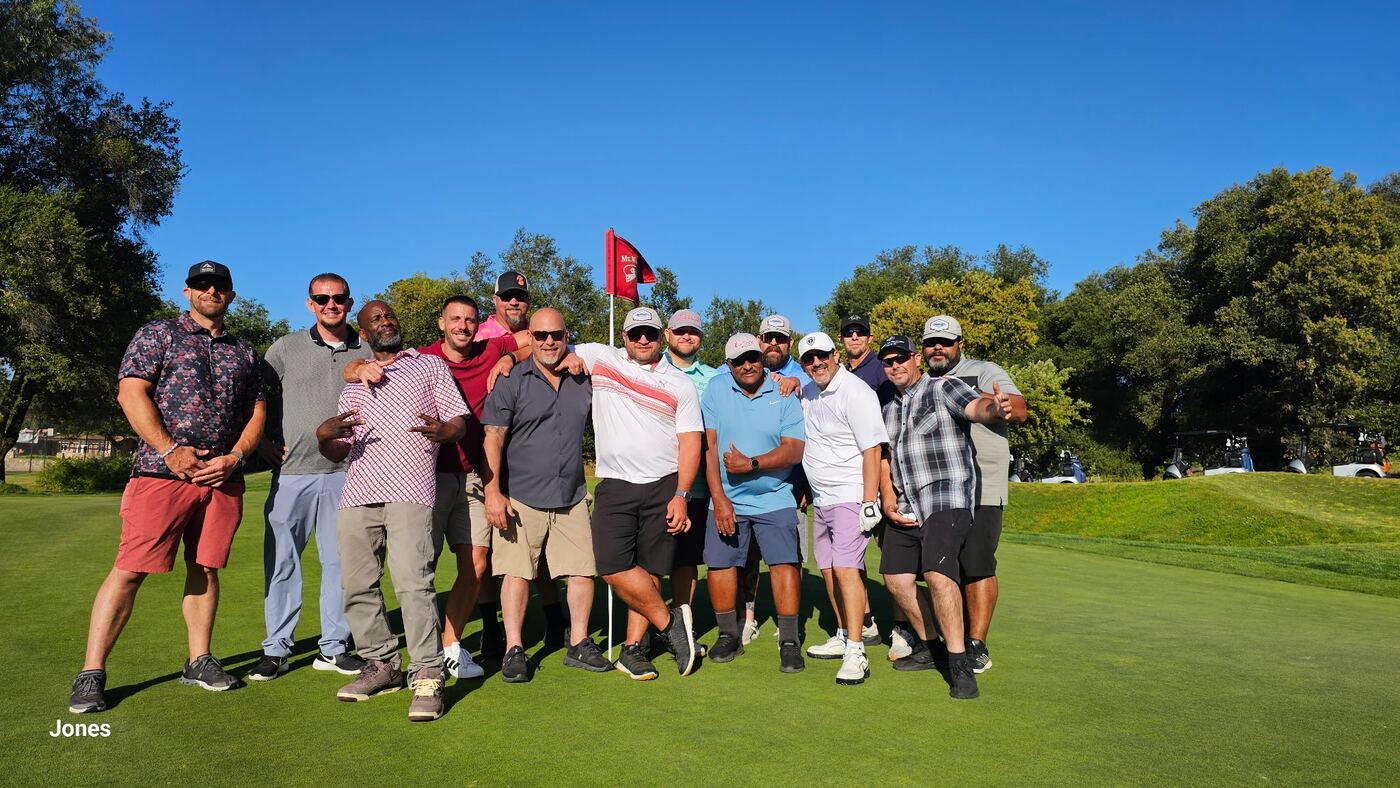 Whole crew on the green at the Jones — flag up, full lineup, perfect bluebird day
