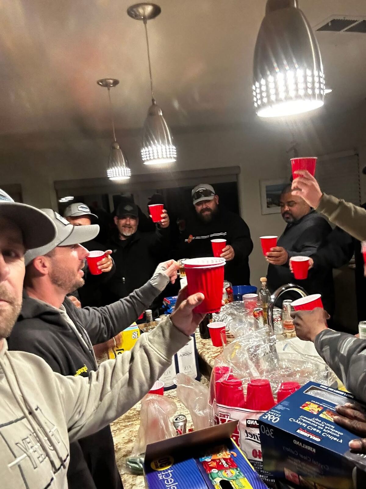 Kitchen cheers under the pendant lights — Solo cups raised, snacks across the counter